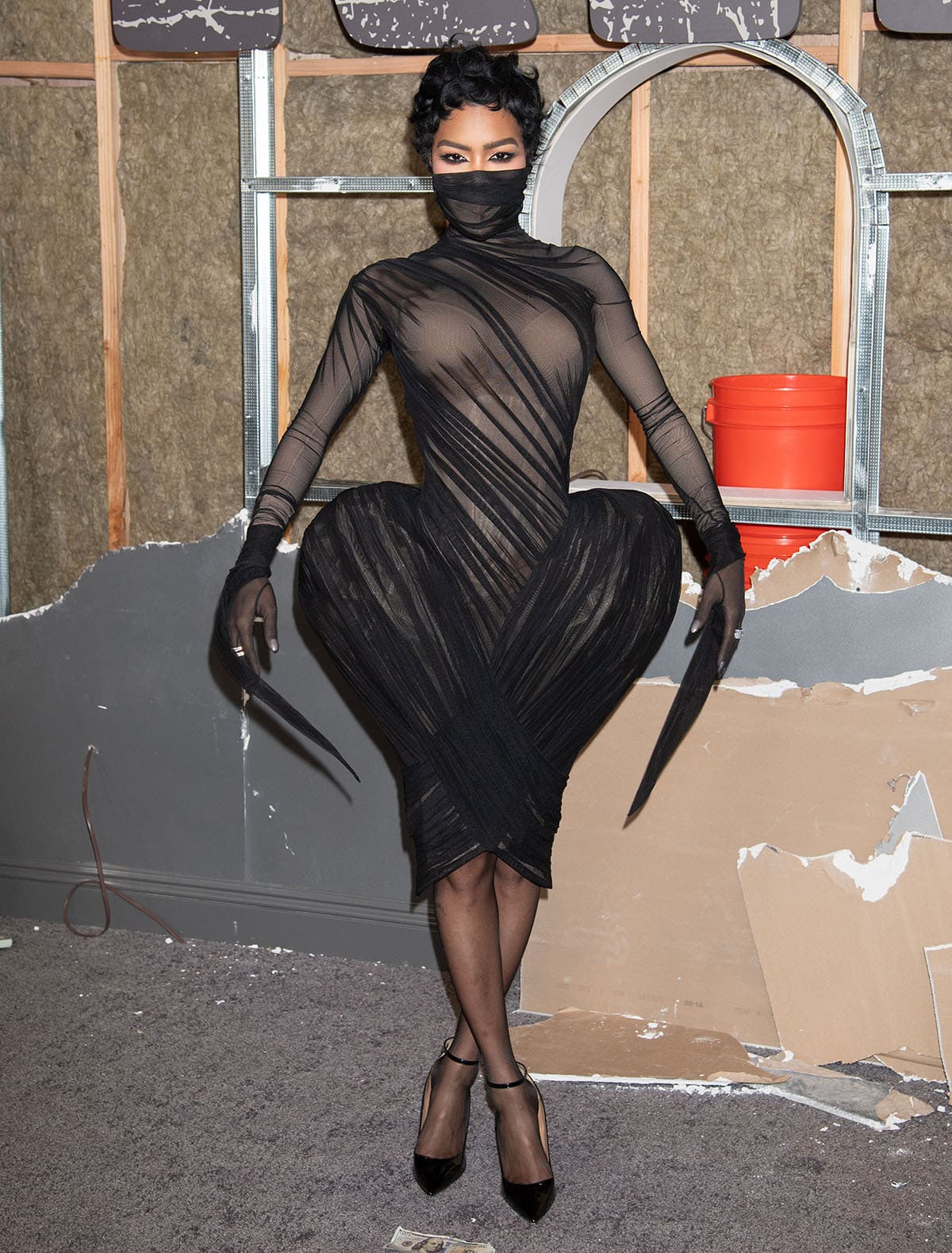 Teyana Taylor commands attention in a sheer black Ashi Studio draped dress featuring an architectural tulip-shaped skirt, long sleeves with extended cuffs, and a high neck that covers her mouth and nose