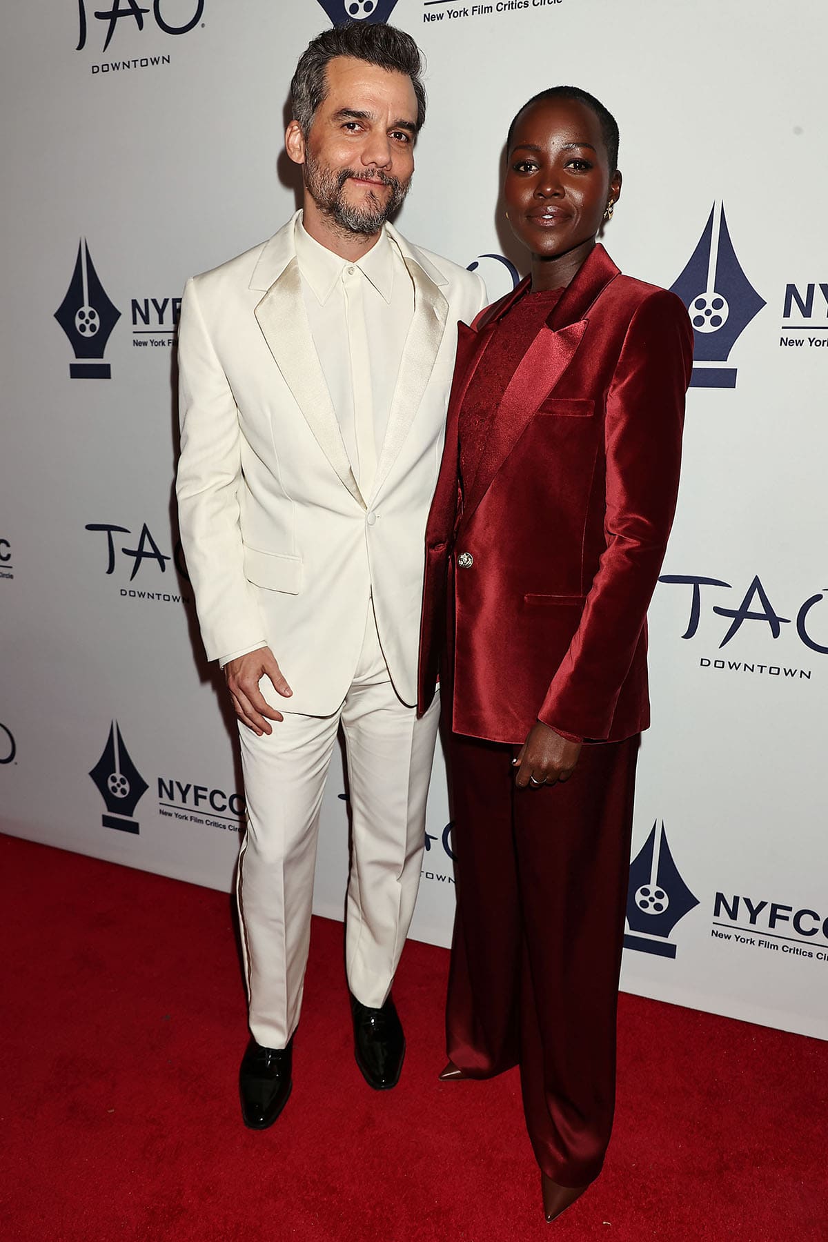 Lupita Nyong’o arrives with Best Actor winner Wagner Moura at the 91st New York Film Critics Circle Awards, wearing a rich red velvet Alice + Olivia suit layered over a sheer lace blouse
