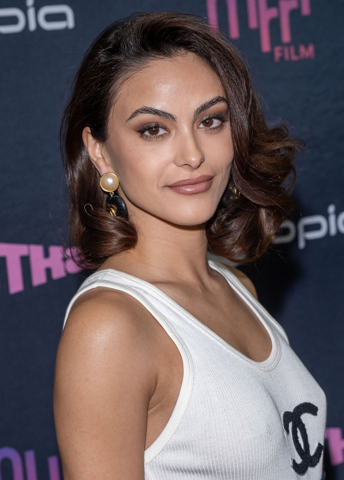 Camila Mendes looks stunning in her retro-inspired side-parted hairstyle, neutral makeup, and statement Chanel earrings featuring large faux pearl studs and dangling black heart charms