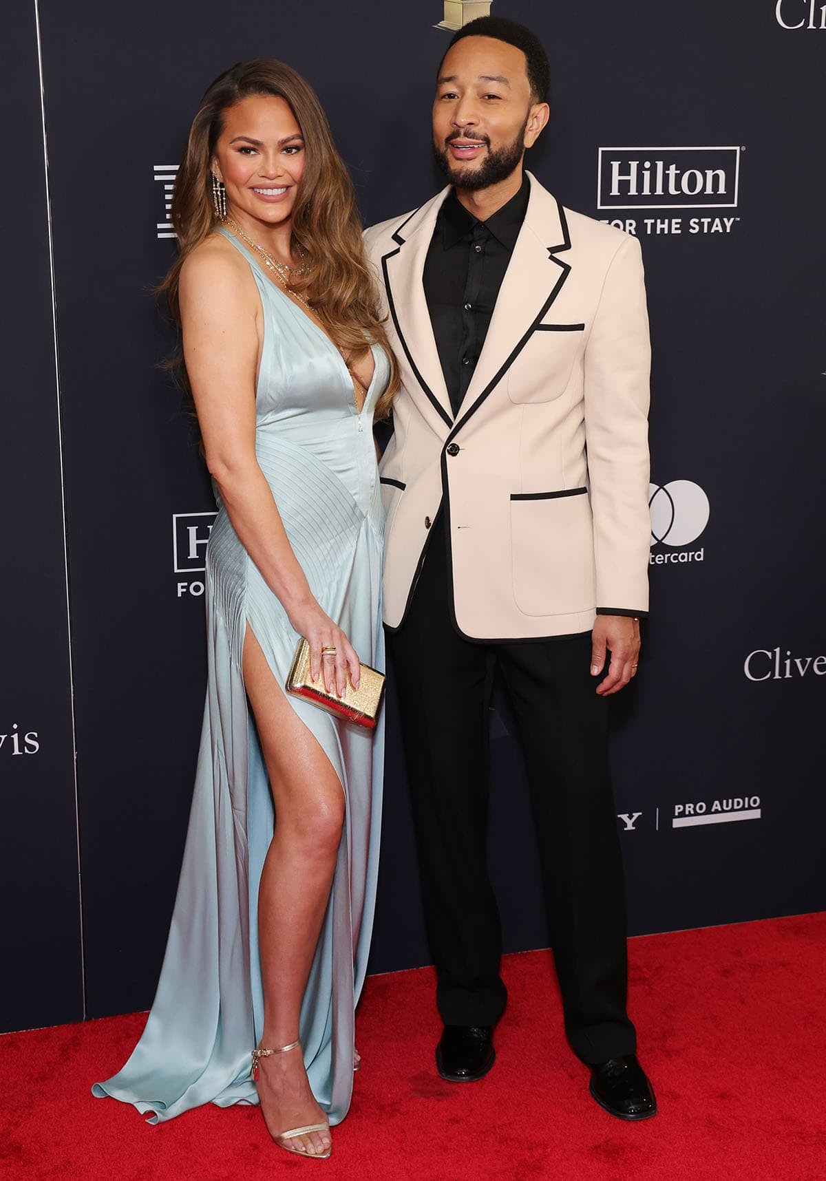 Chrissy Teigen looks stunning in her aqua blue Tom Ford gown as she poses beside her husband, John Legend, who looks dapper in a white Valentino blazer with black piping, black trousers, and dress shoes