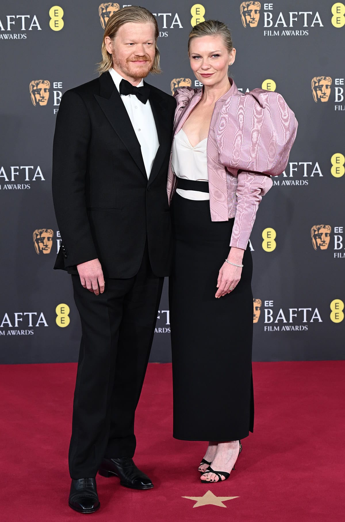 Kirsten Dunst, with her husband Jesse Plemons, power dresses in a Valentino ensemble featuring a white blouse, a black skirt, and a pink moiré jacket with oversized mutton sleeves