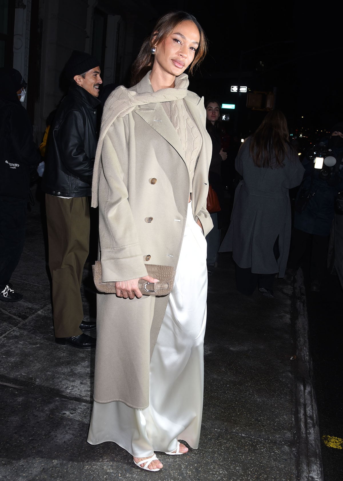 Joan Smalls elevates relaxed winter neutrals by pairing delicate white heels with fluid satin trousers, a tan cable-knit top, and a softly tailored beige trench for an effortlessly polished finish