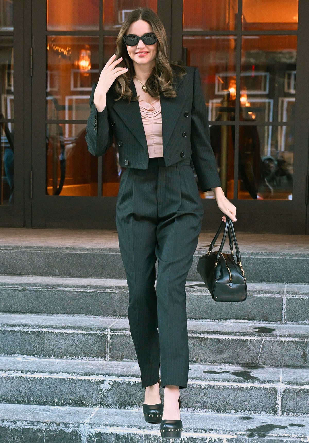 Miranda Kerr pairs her clog-like studded pumps with a black pinstriped suit, featuring a cropped blazer, high-waisted tapered pants, and a pink cropped top with a gathered neckline