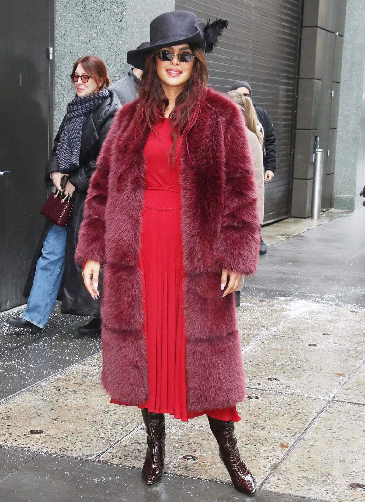 Priyanka Chopra channels piratecore energy by pairing her burgundy knee-high boots with a red jersey dress, a burgundy Apparis “Sykle Sable” faux-fur coat, and a black cavalier hat with a feather accent