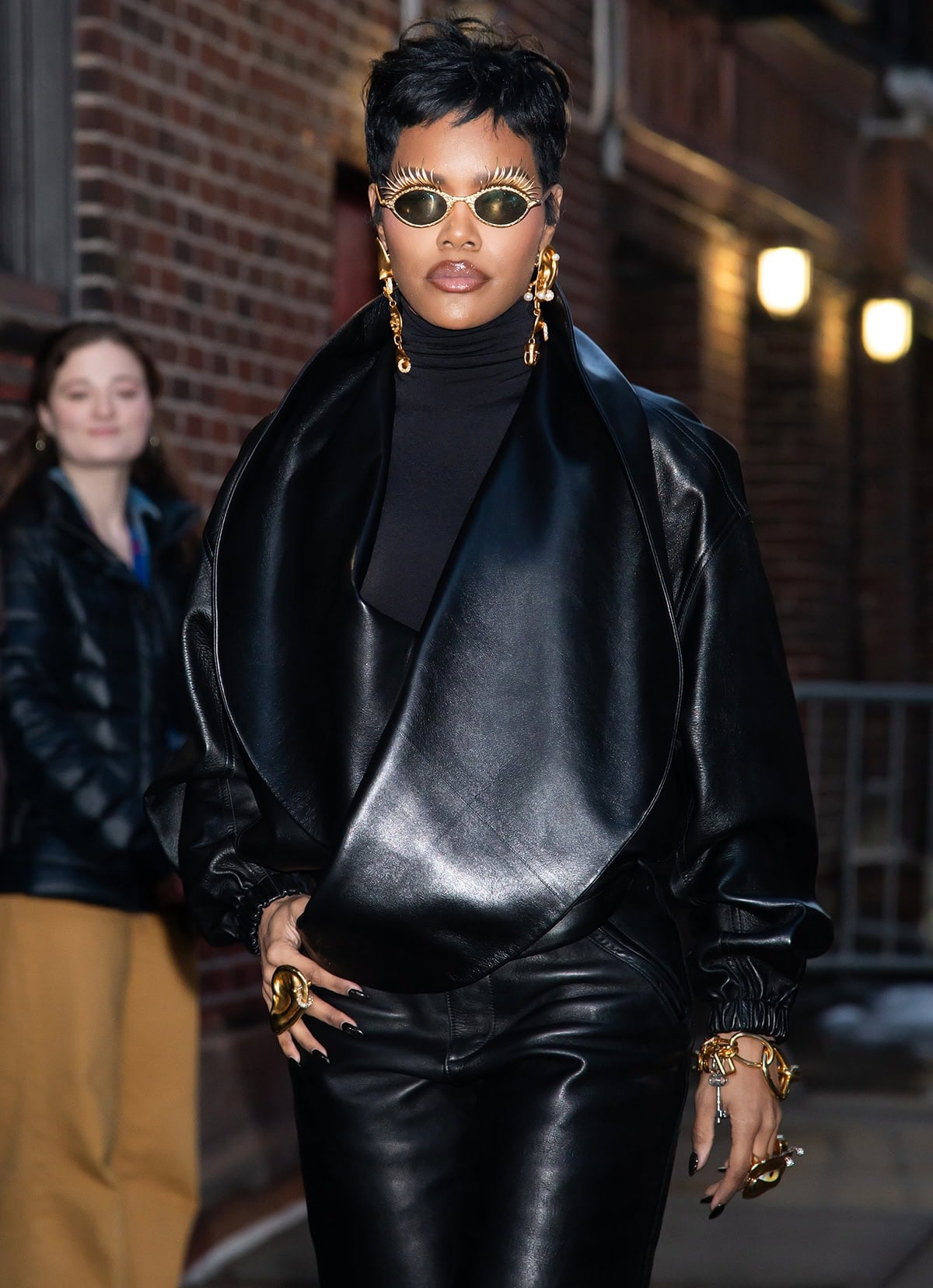 Teyana Taylor adds a touch of whimsy to her tough-chic all-black leather look with gold jewels by Schiaparelli, including a pair of hammered sunglasses with eyelashes