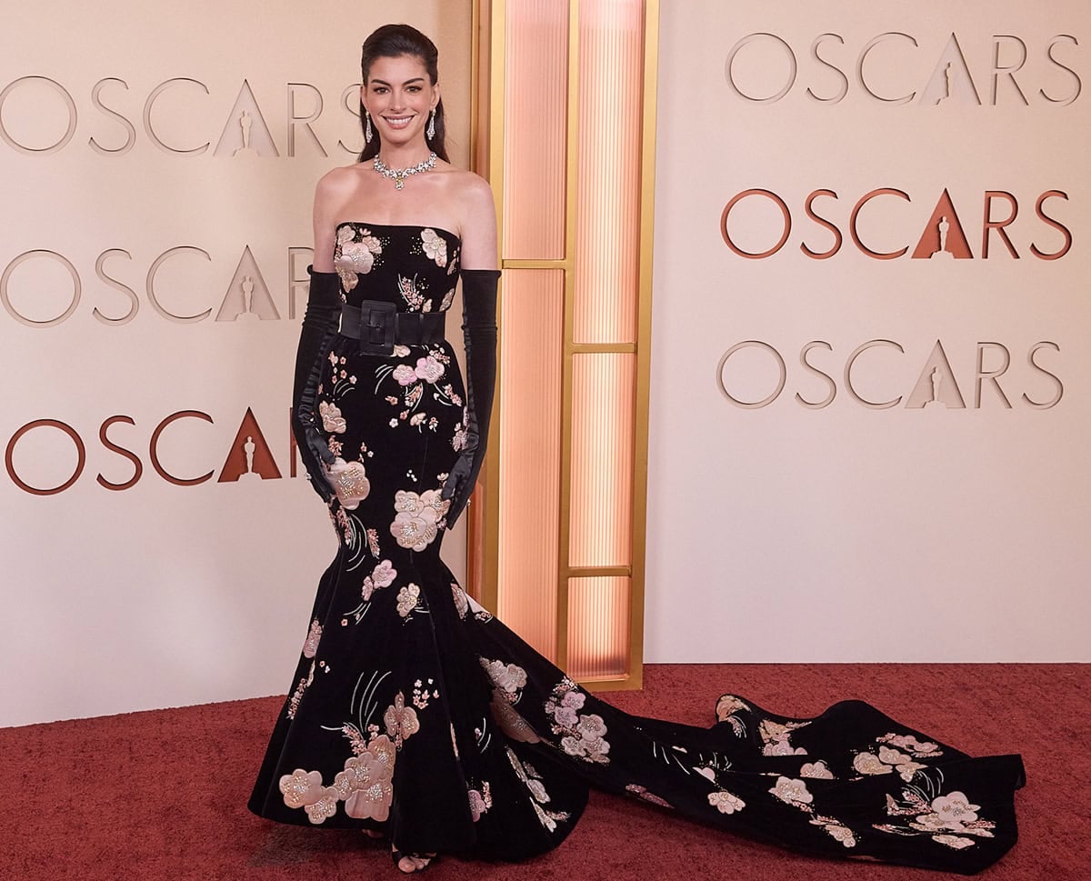 Anne Hathaway stuns in a custom black Valentino floral-embroidered gown with a belted waist, a mermaid skirt, a sweeping train, and black opera gloves