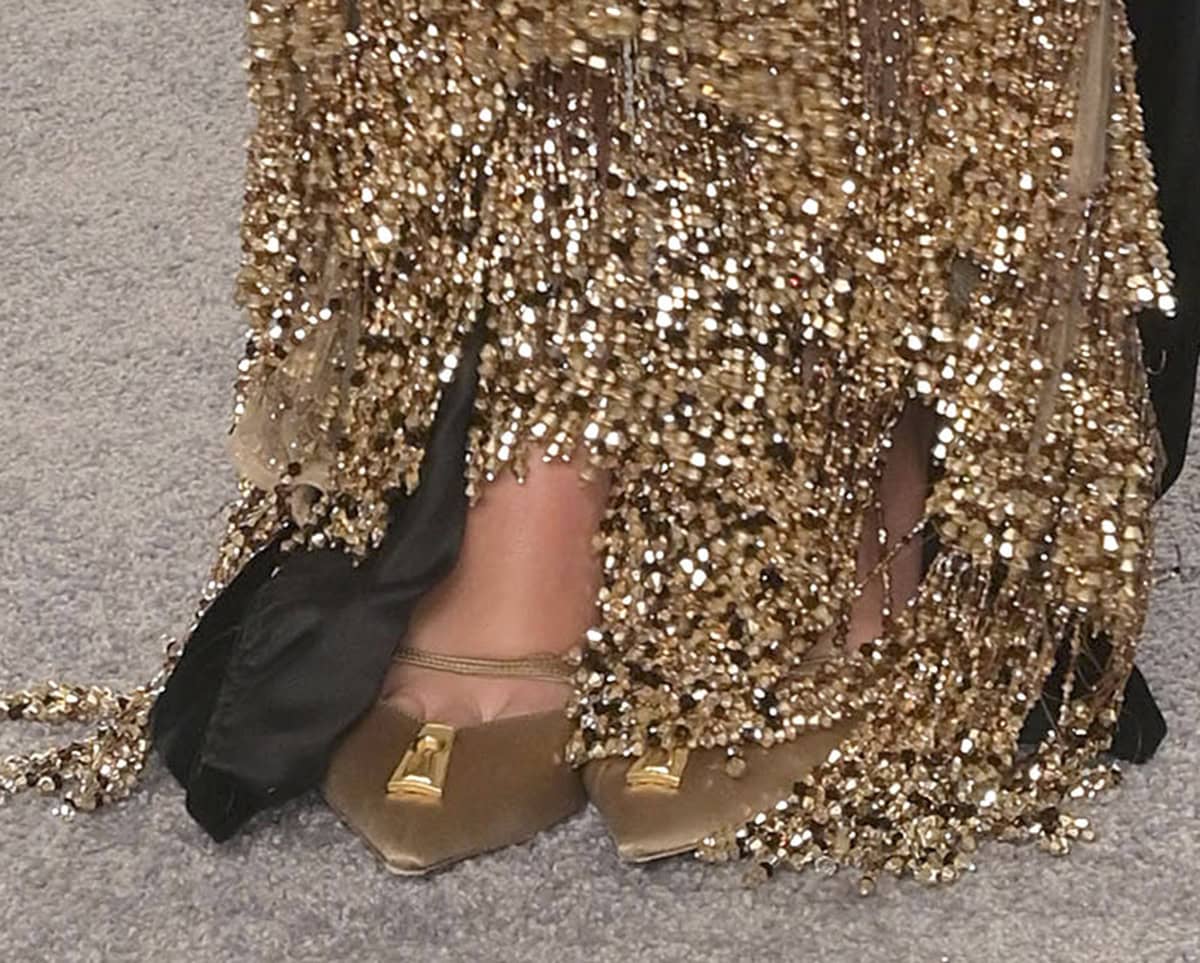 Dua Lipa parties at the Vanity Fair Oscars celebration in a pair of gold satin pumps by Schiaparelli, featuring a keyhole motif, a slim strap, and low heels