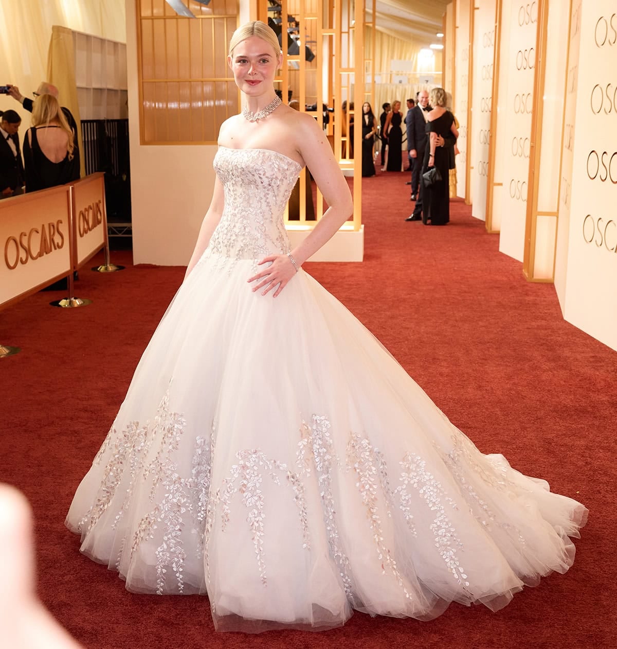 Elle Fanning shows her signature whimsical style in a custom white Givenchy gown with a bustier bodice and a voluminous tulle skirt, both with hand-stitched silver satin wisteria