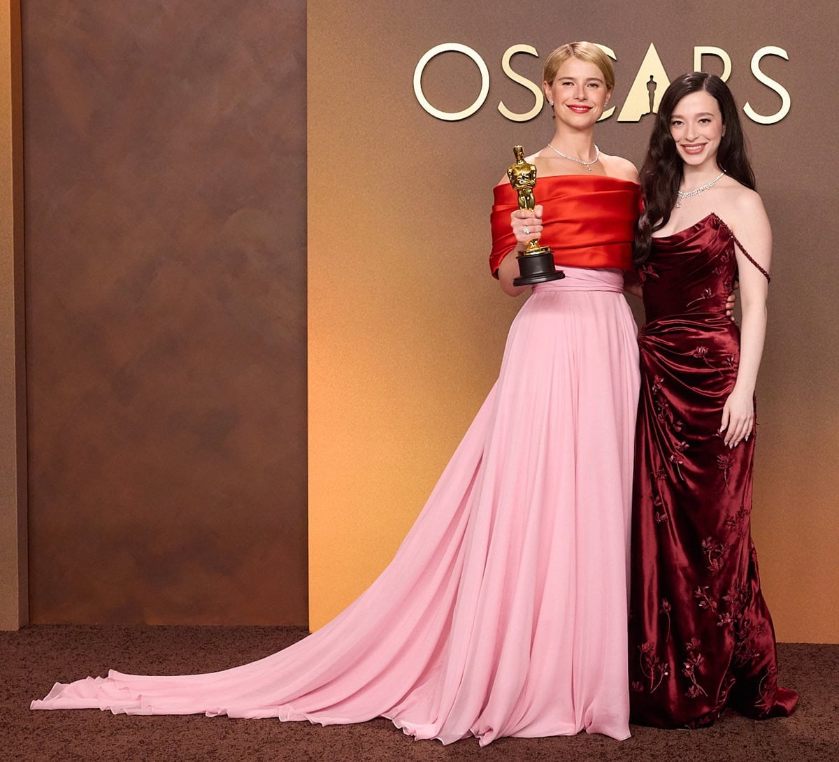 Jessie Buckley poses with her Oscar for Actress in a Leading Role beside Mikey Madison, who stuns in a rich red velvet Dior gown with floral appliqués, a thigh-high slit, and delicate beaded off-the-shoulder straps, styled with Tiffany & Co. jewelry and glossy Old Hollywood waves