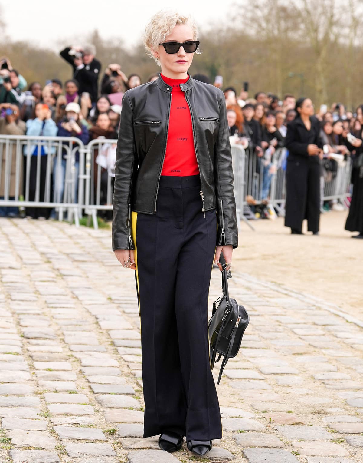 Julia Garner pairs her architectural Loewe pumps with a black leather moto jacket, a red turtleneck top, and wide-leg pants with yellow side stripes