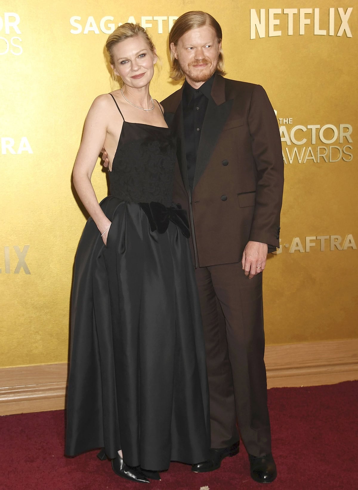 Kirsten Dunst, with her husband Jesse Plemons, embraces an edgy, feminine style, pairing her black shoes with a black Khaite dress featuring a square neckline, floral lace embroidery on the bodice, and a black velvet bow at the waist