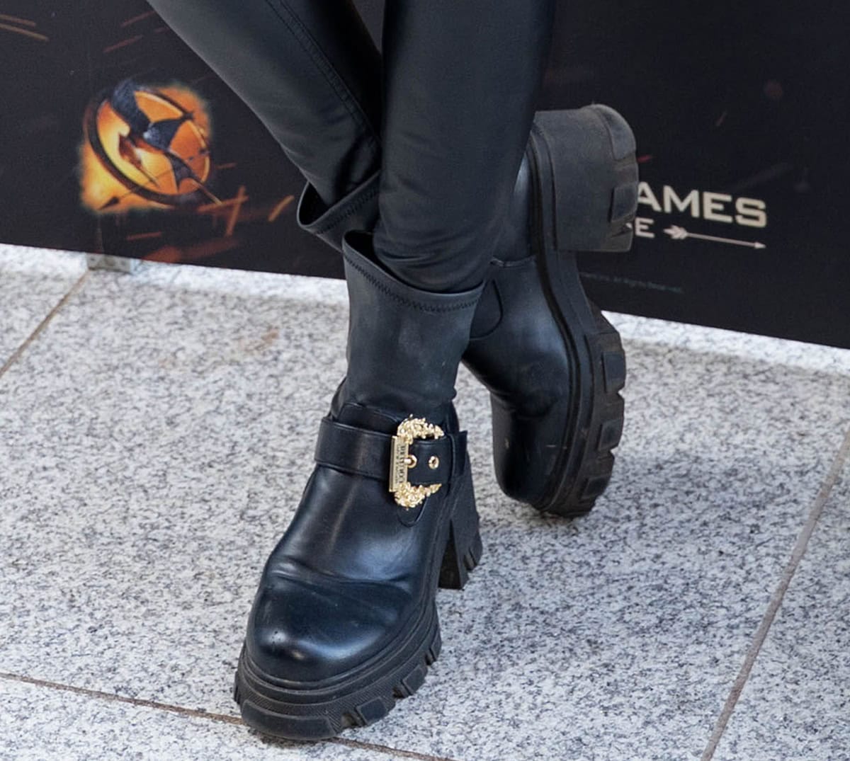 Laura Whitmore wears the black leather Versace “Sophie” ankle boots featuring a large gold-tone baroque buckle, platforms, and short block heels