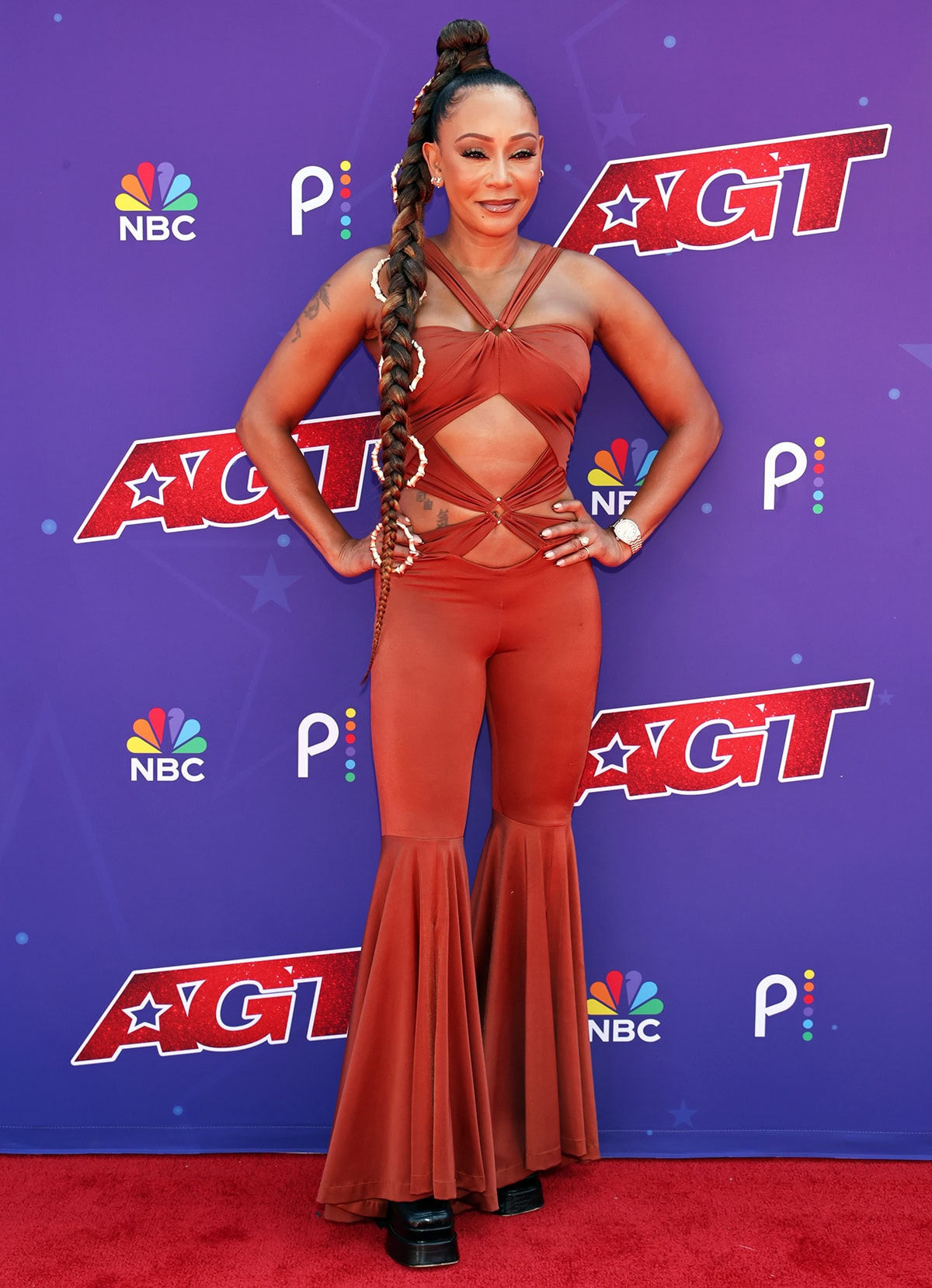 Mel B pairs her striking black boots with a copper jumpsuit by Maria Kalkoff featuring a halter strap, cutout panels with gathered straps and silver-tone rings, and softly pleated pants