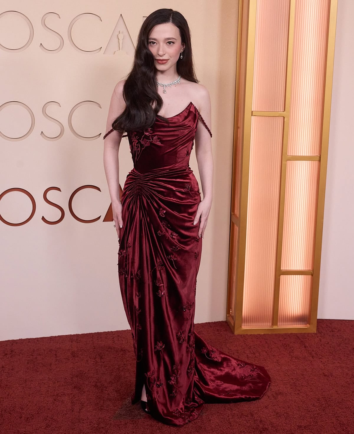 Mikey Madison looks stunning, pairing her black heels with a custom red velvet Dior gown featuring off-the-shoulder straps, draped skirt, and 3D flower appliques