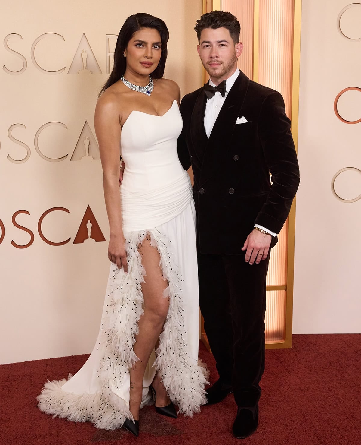 Priyanka Chopra stuns in a white feather-trim Dior gown styled with black Jimmy Choo “Brigitte” satin pumps while posing with Nick Jonas at the 98th Academy Awards