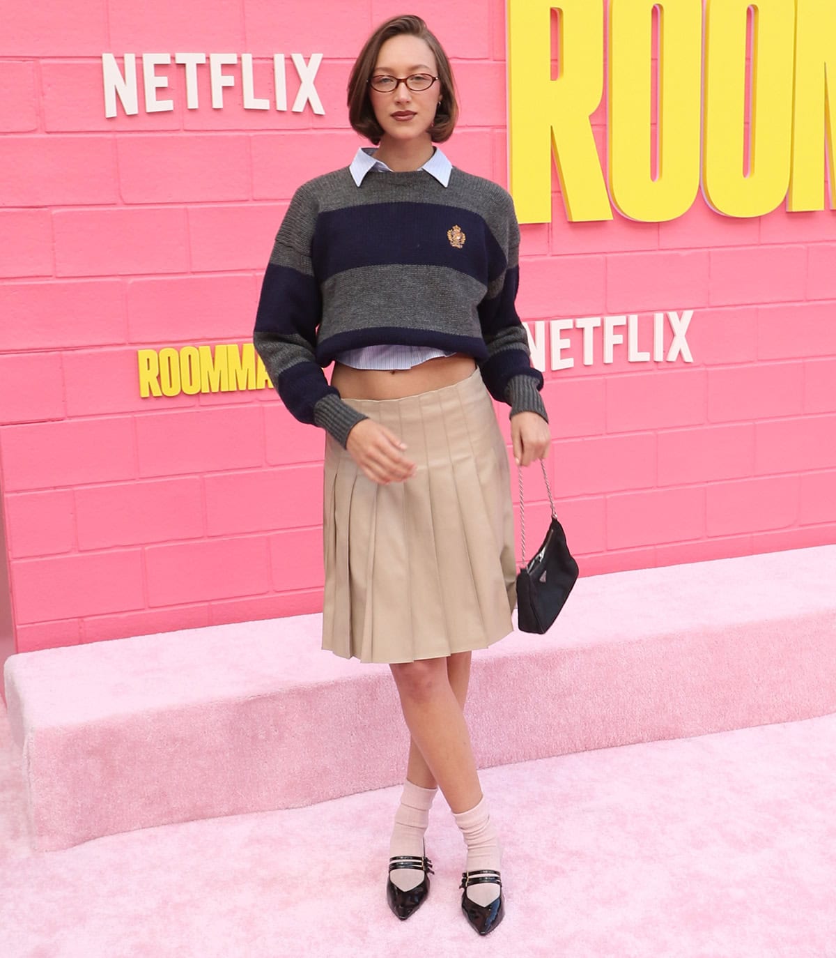 Ava Michelle pairs her black Mary Janes with thick socks, a pleated beige knee-length skirt, a collegiate-style sweater, and a cropped shirt