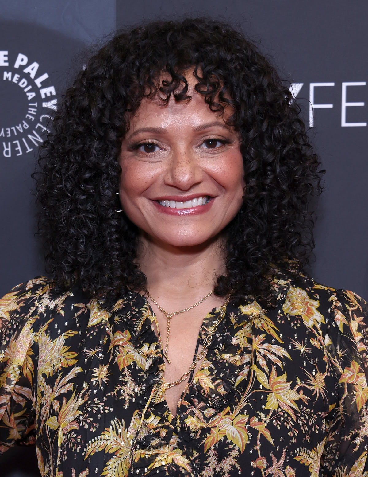 Judy Reyes styles her look with hoop earrings, carries a Loeffler Randall “Bailey” clutch in brown-and-yellow striped raffia, wears her hair in tight curls, and layers two chain necklaces around her neck