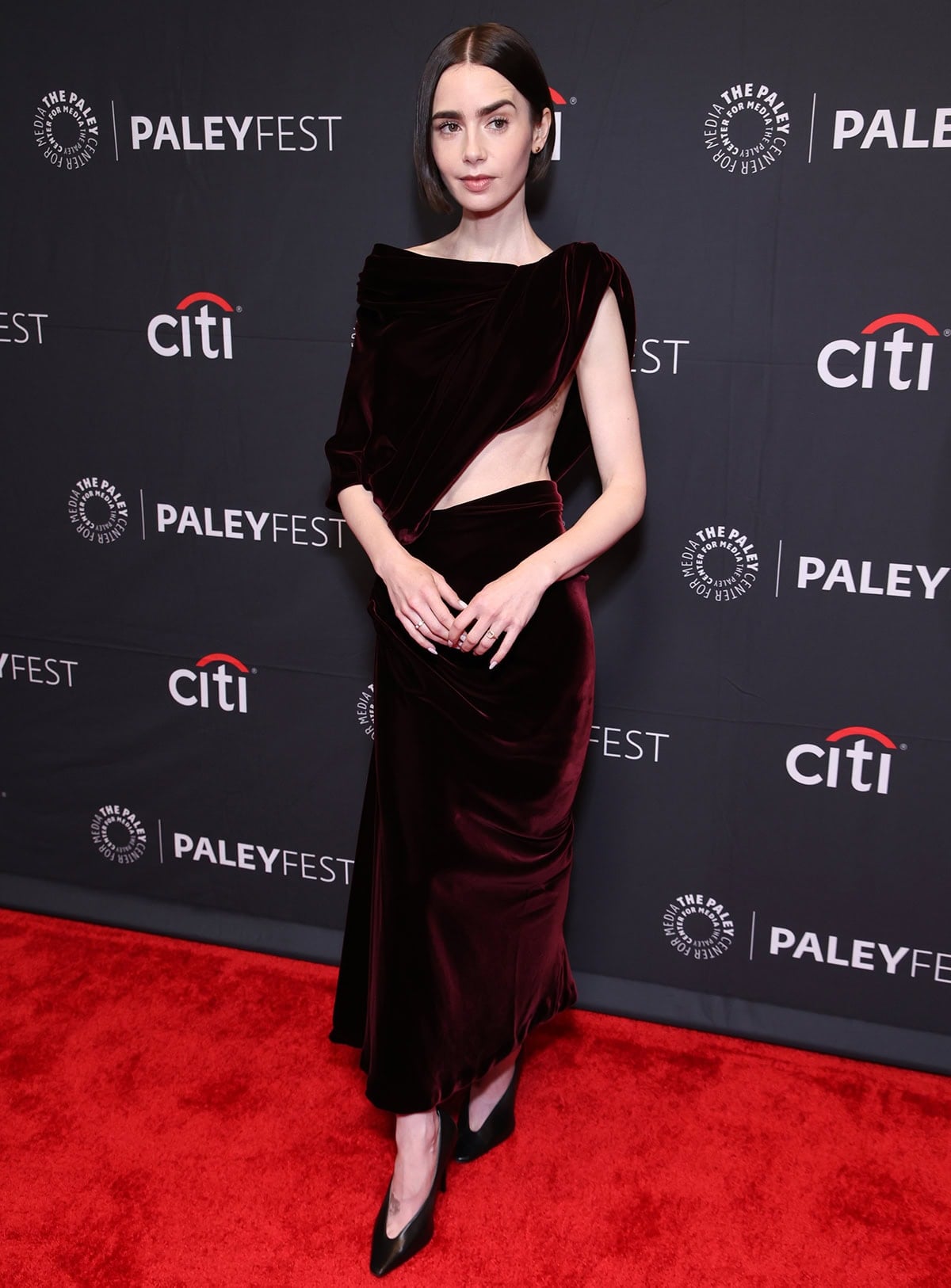 Lily Collins teams her sleek, modern pumps with a rich red velvet draped dress with a bold asymmetrical side cutout by Balenciaga