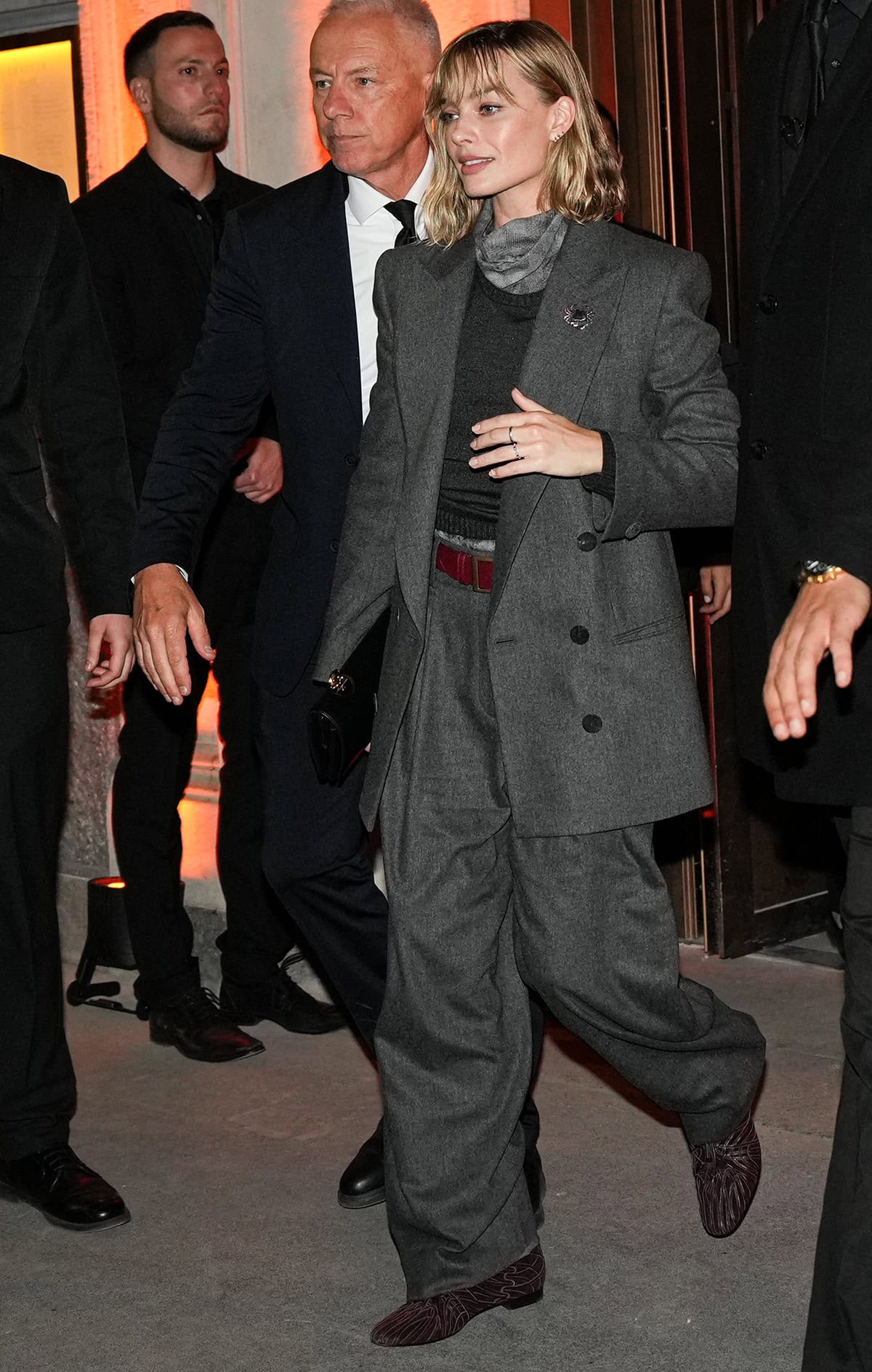 Margot Robbie pairs her burgundy oxfords with a charcoal gray Armani suit, a gray knit top, a printed high-neck blouse, and a red belt