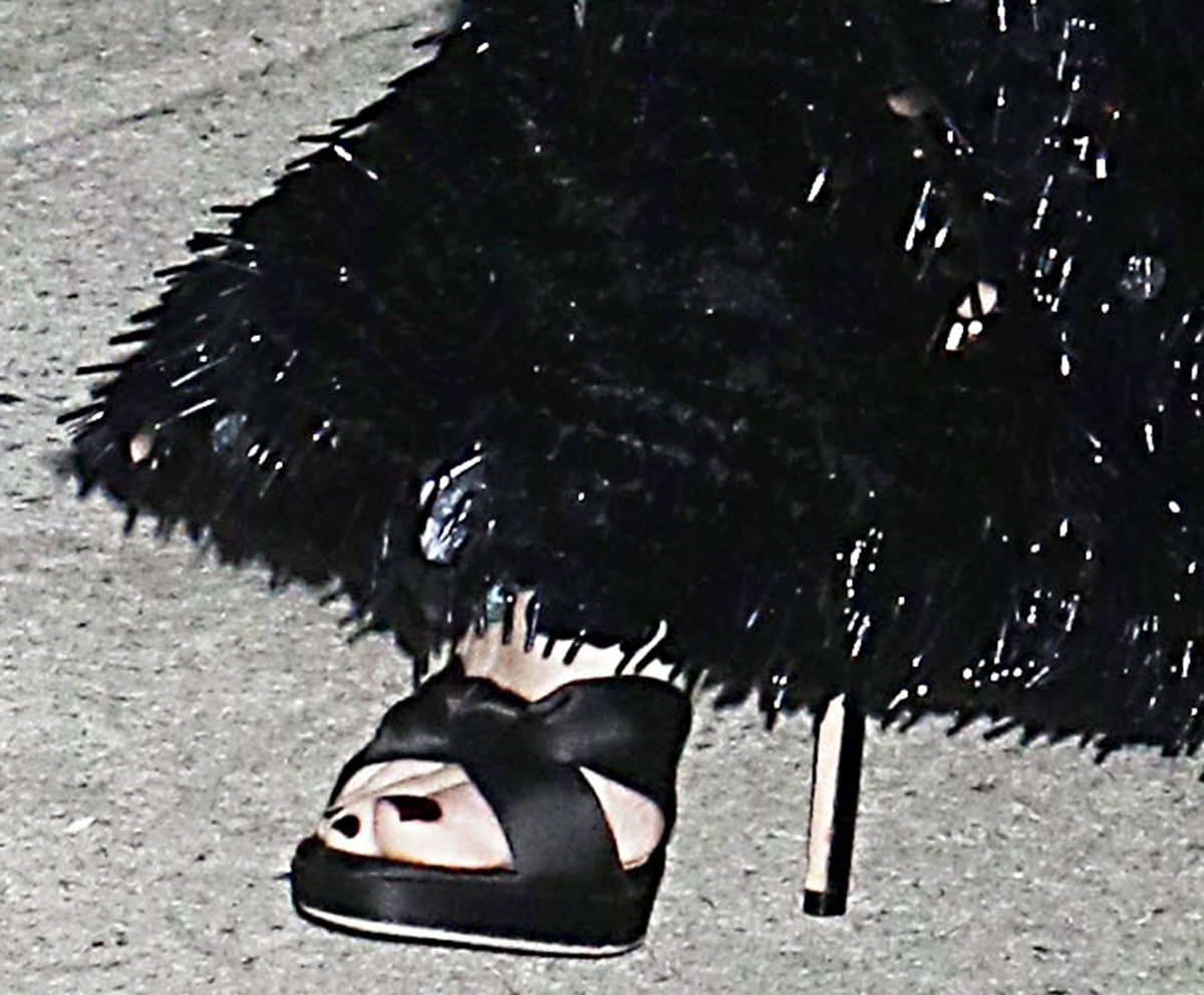 Naomi Watts slips her black-pedicured feet into the Jimmy Choo “Rosie” sandals featuring knotted front straps, delicate ankle straps, platforms, and stiletto heels