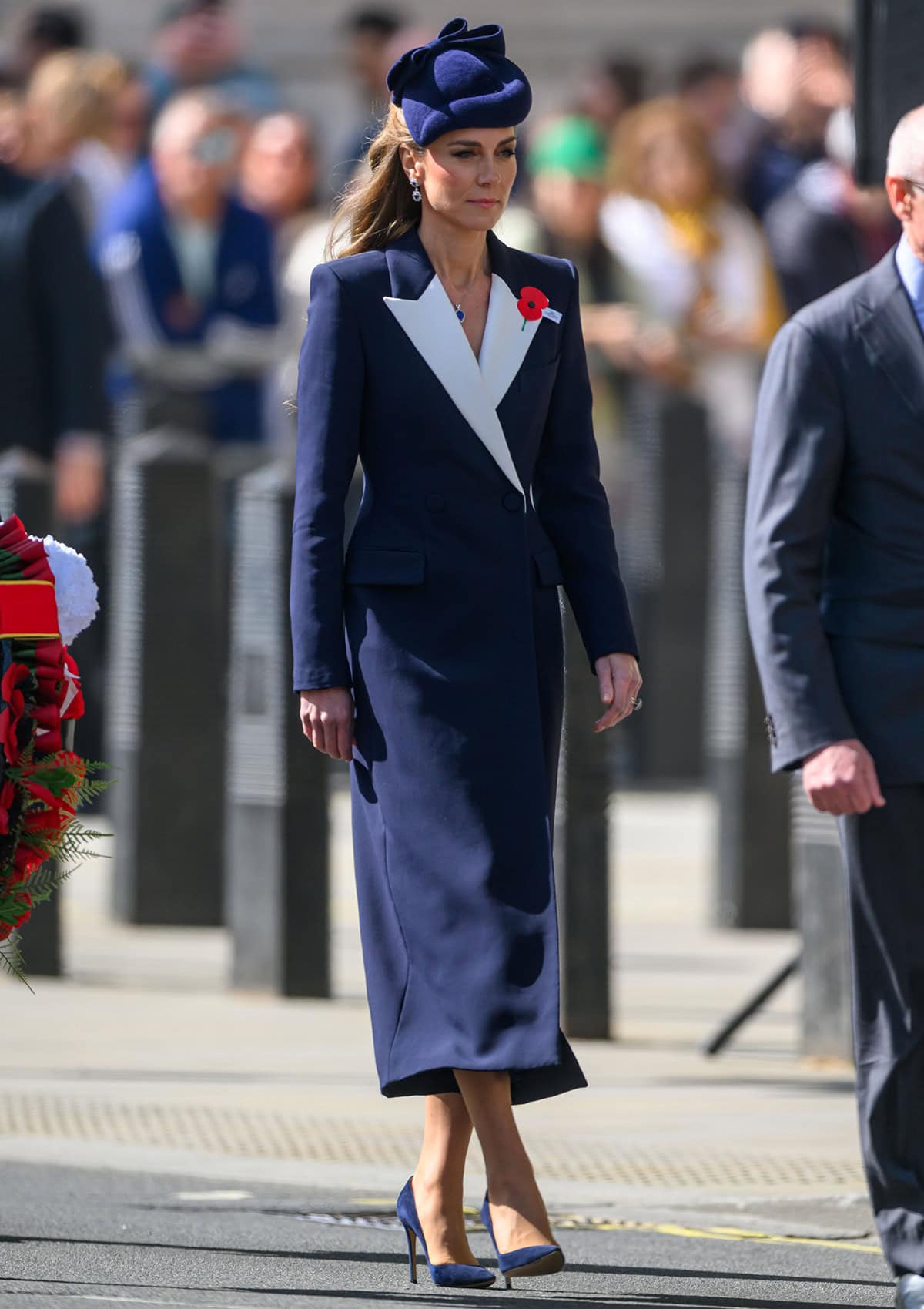 Kate Middleton pairs her navy blue pumps with a matching double-breasted navy coat dress by Givenchy featuring contrasting white lapels, a red poppy flower accent, and padded shoulders