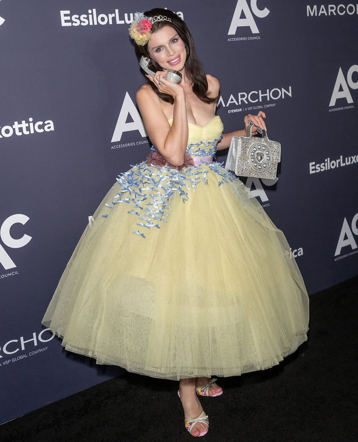 Julia Fox pairs her feminine floral mules with a vintage yellow Betsey Johnson gown featuring a gathered sweetheart neckline, tiny blue ribbons, a pink sash belt with plum floral appliques, and a full, voluminous skirt