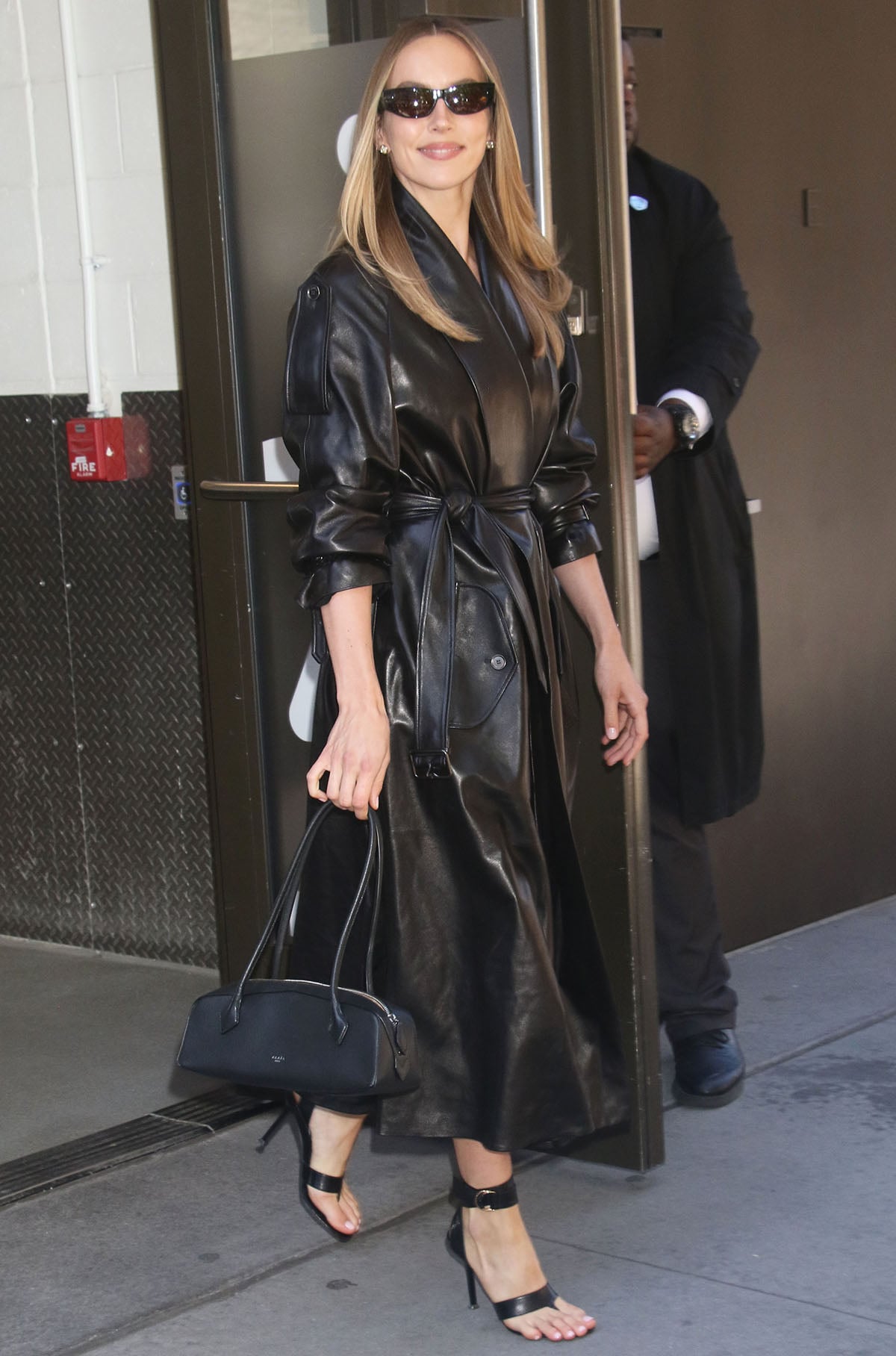Jodie Comer styles Christen's architectural "Helix" sandals with a belted black Burberry leather coatdress, Alaïa's Le Teckel bag, and slim sunglasses for a sleek monochromatic look