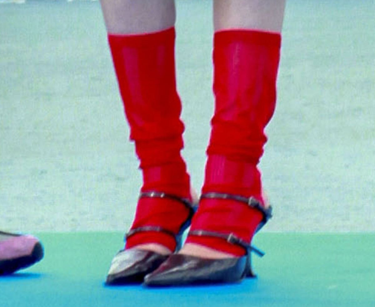 Milly Alcock wears red leg warmers with the Miista “Shari” mules in black leather with pointed toes, two thin buckled straps across the foot, and high heels with an angular base
