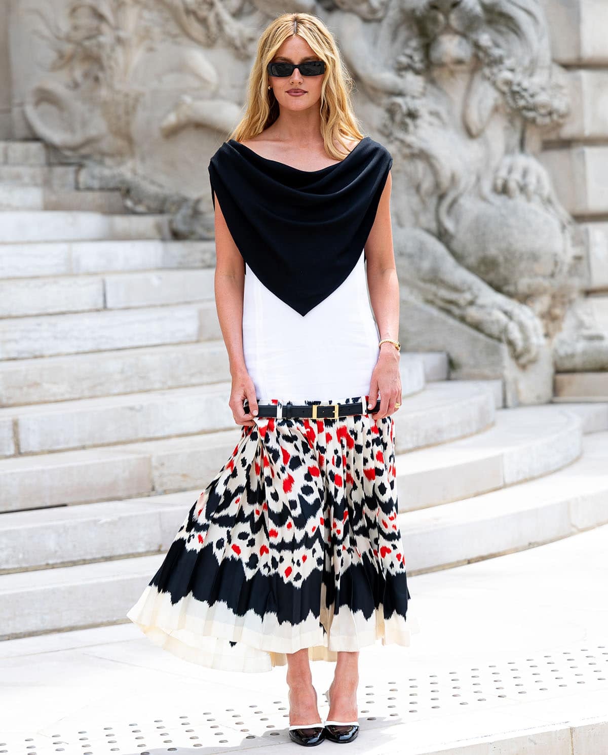 Sarah Pidgeon pairs her glossy two-tone slingback pumps with a Chanel sleeveless dress featuring an integrated black scarf-like bib, a belted drop waist, and a painterly pleated midi skirt